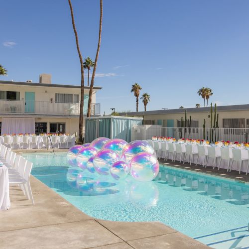 An outdoor pool scene with tables set for an event, featuring vibrant flowers and floating translucent beach balls under clear skies.