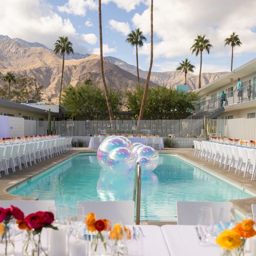 Outdoor dining setup by a pool with balloons, palm trees, and mountains in the background. Tables have floral centerpieces.