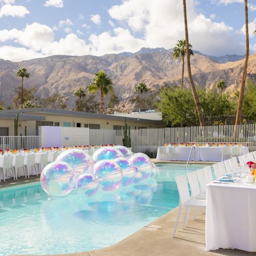 A poolside setup with long dining tables and floral centerpieces, surrounded by mountains and palm trees, features floating transparent balloons.