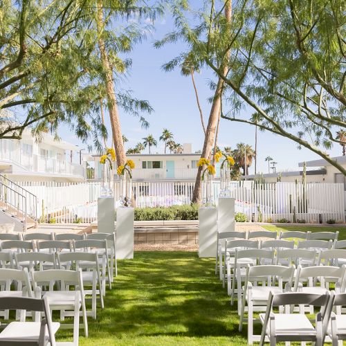 An outdoor wedding setup with white chairs arranged in rows on a lawn, flanked by trees, leading to a small altar area.