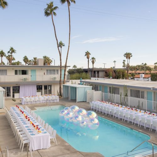 Outdoor pool with floating decorations, flanked by long banquet tables. Modern buildings and palm trees surround the venue.