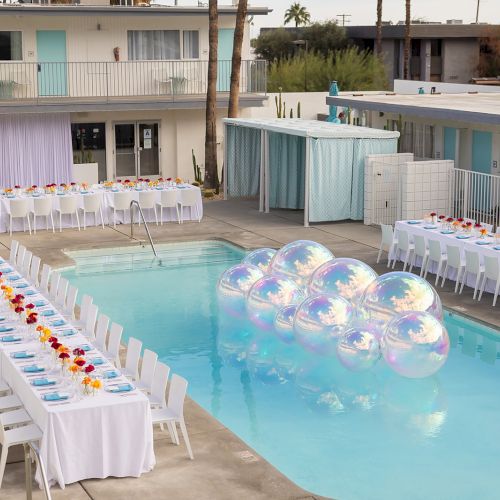 Poolside setup with long tables, white chairs, and colorful plates for an outdoor event. Large iridescent balloons float in the pool.