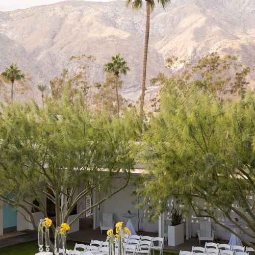 Outdoor wedding setup with white chairs and floral arrangements, set amidst trees and mountains under a clear sky.