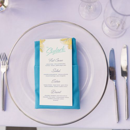 A dining setup with a menu placed on a blue napkin, surrounded by a plate, cutlery, and glasses on a white tablecloth.