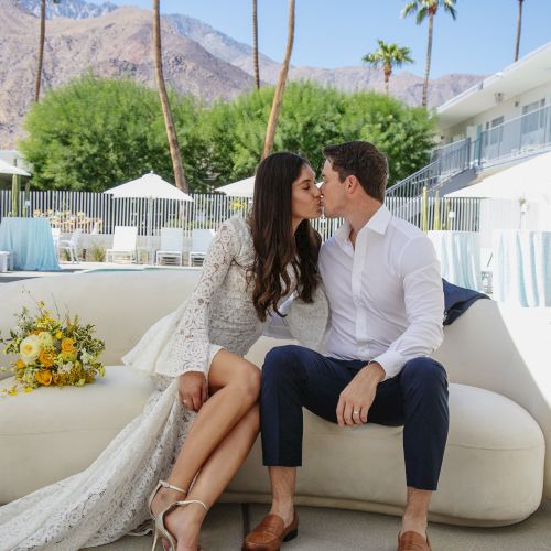 A couple dressed in semi-formal attire share a kiss on a couch at an outdoor venue, surrounded by white furniture and palm trees with mountains in the background.