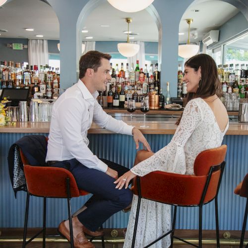 A couple sits at a bar, smiling at each other. The bar has a bouquet of yellow flowers and a modern design, with bottles and mirrors behind them.