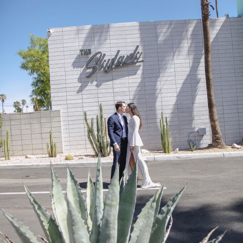 A couple stands in front of a building with the word "Skylands" on it, surrounded by desert plants and a clear blue sky.