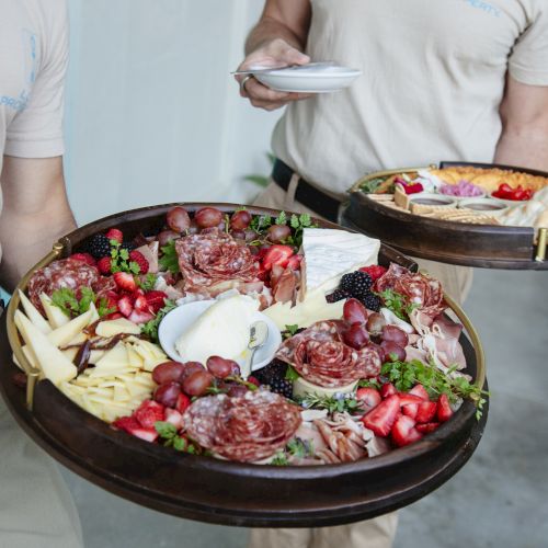 Two people are holding trays with an assortment of cold cuts, cheeses, fruits, and other hors d'oeuvres. The trays are artistically arranged.