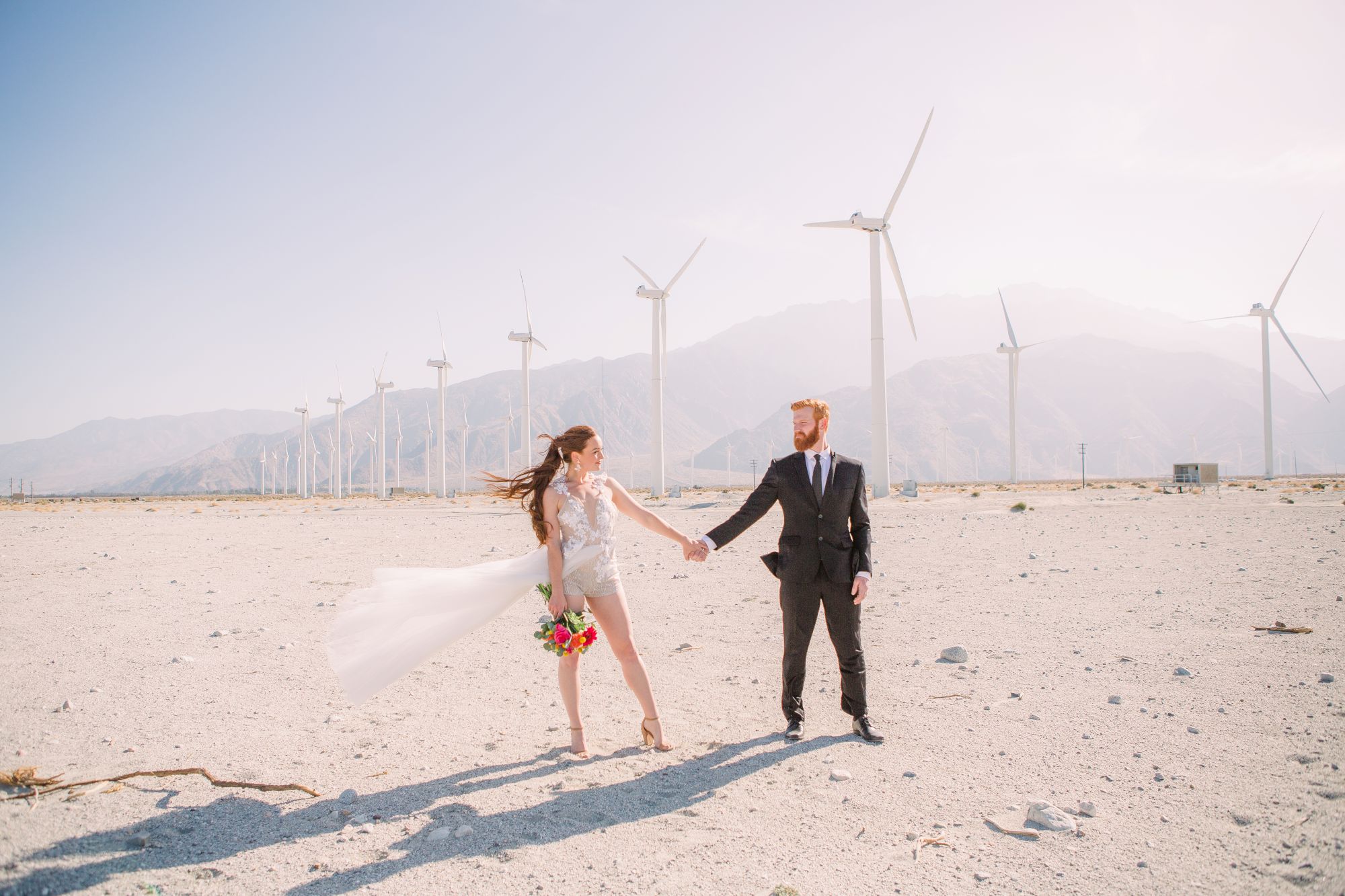 A couple in wedding attire holds hands in a desert landscape with wind turbines in the background, under a clear blue sky.