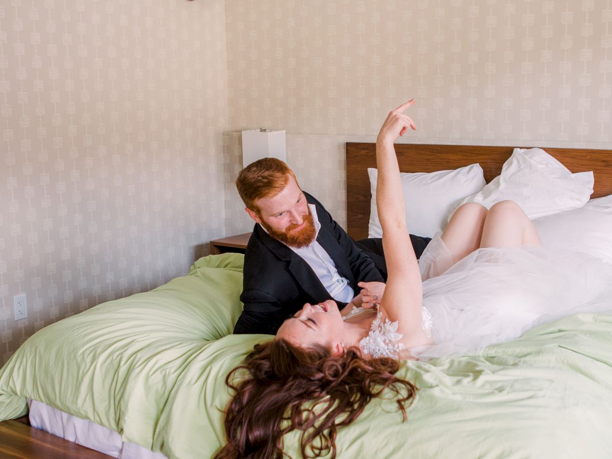 A couple is lying on a bed in a cozy room with floral decor, where one person is playfully reaching up, and a harness is mounted on the ceiling.