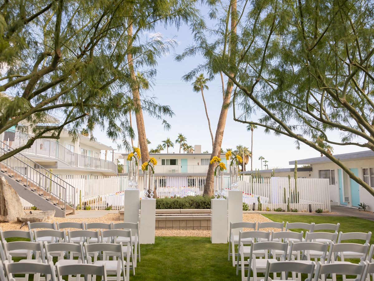 An outdoor wedding setup with white chairs, floral decorations, and greenery, surrounded by trees. A serene, festive atmosphere.