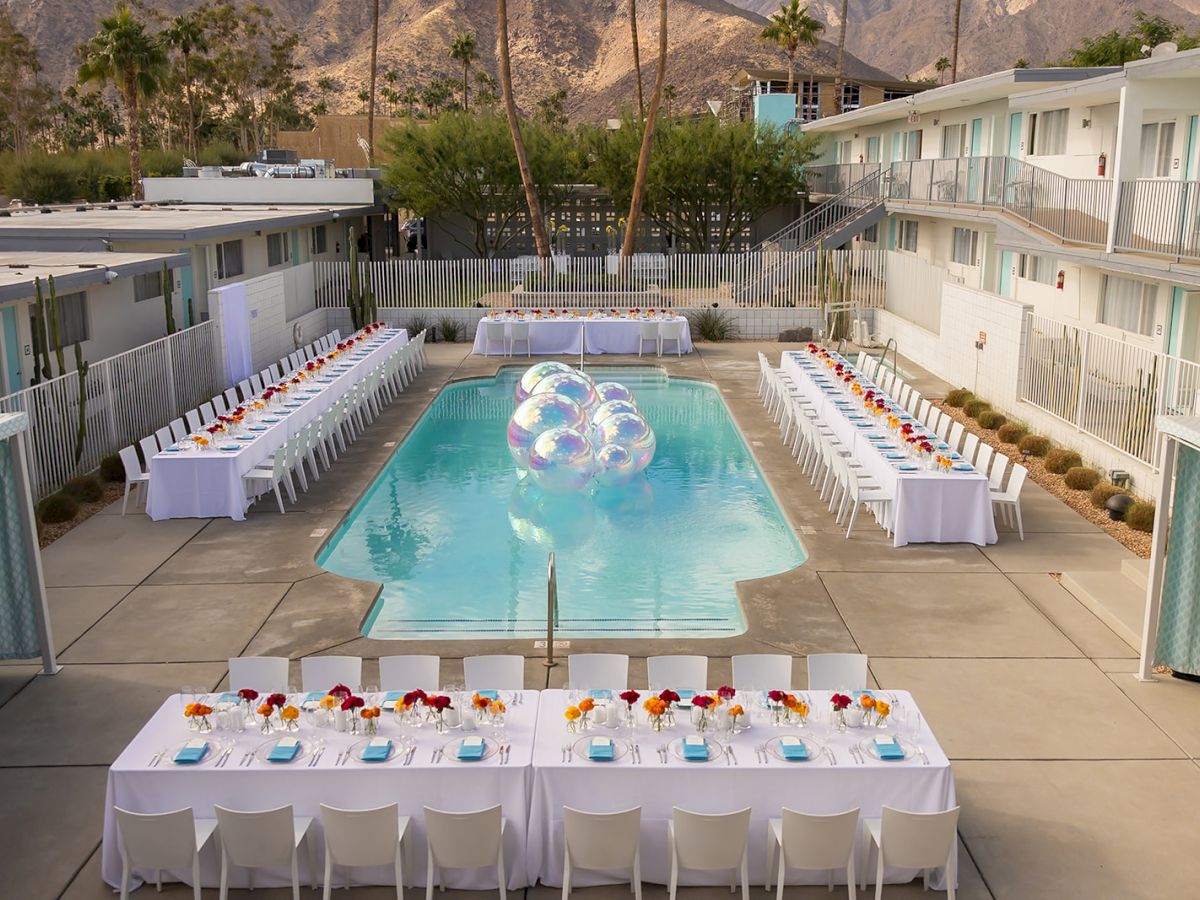 An outdoor setting with a pool, surrounded by long tables set for an event, featuring clear decorative spheres in the water.