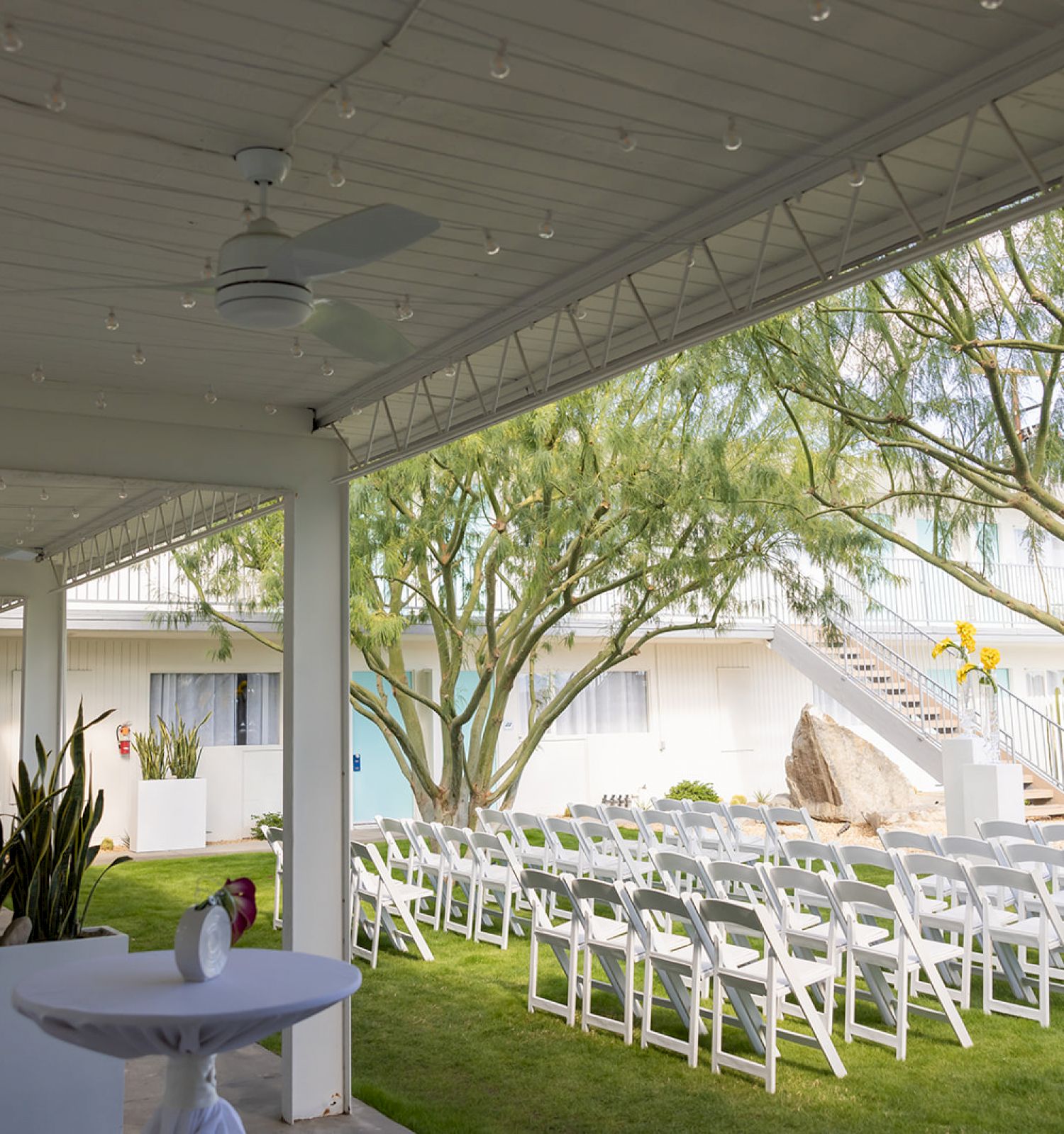 The image shows an outdoor event space with white chairs arranged in rows, tables with white cloths, and greenery, likely set for a wedding.