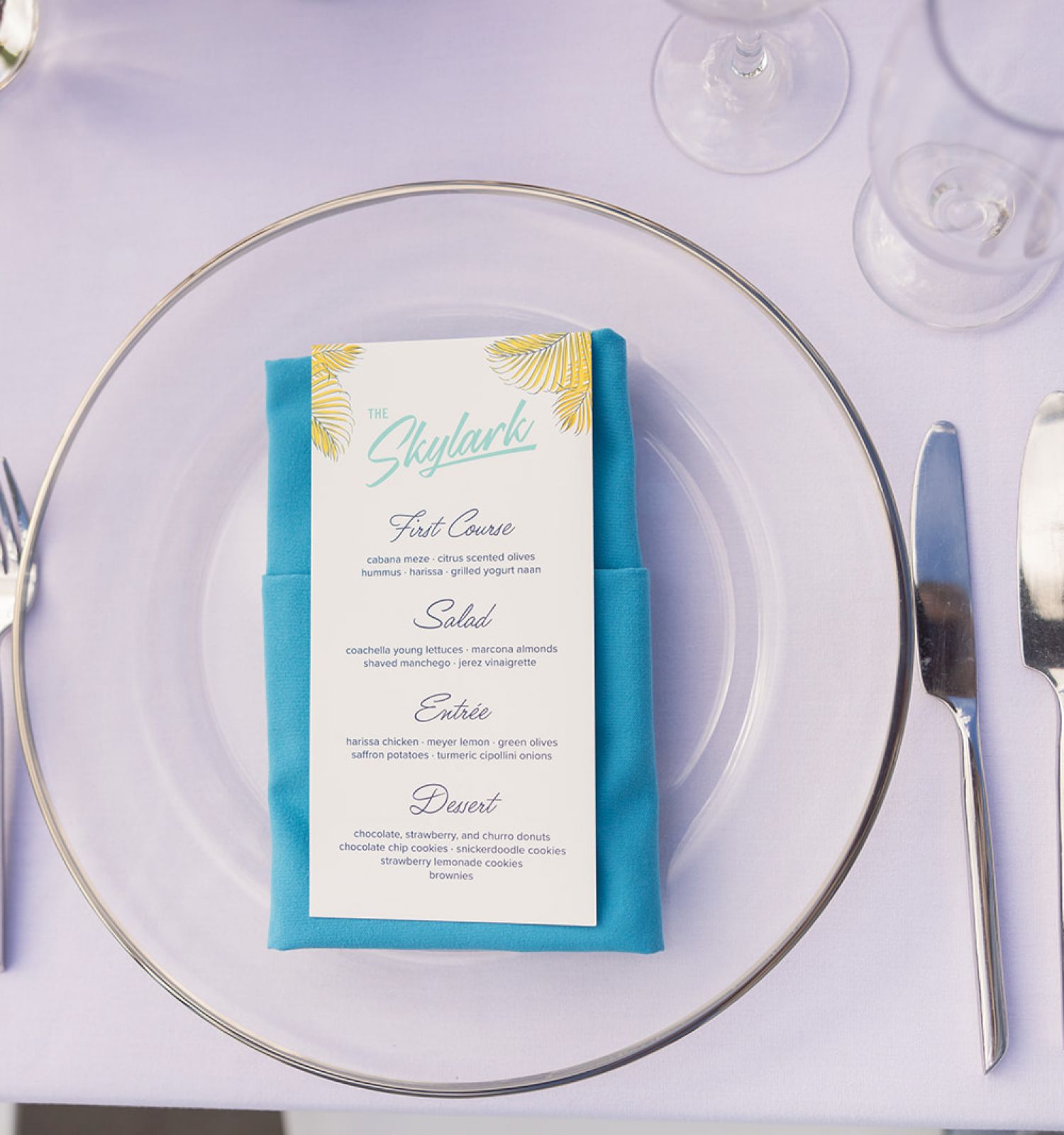 A table setting with a menu in a blue napkin on a clear plate, surrounded by silverware and glasses on a white tablecloth.