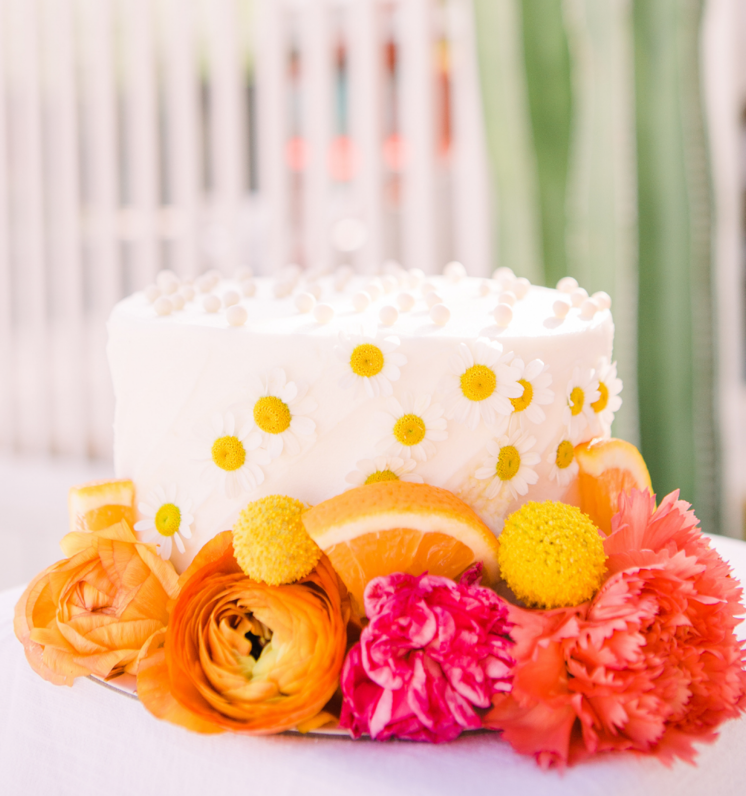 A decorated cake topped with white frosting and yellow dots, surrounded by colorful flowers and orange slices, is set on a white surface outdoors.