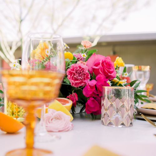 A beautifully set table with an orange glass, floral centerpiece, and decorative candle holders, featuring a vibrant and elegant arrangement.