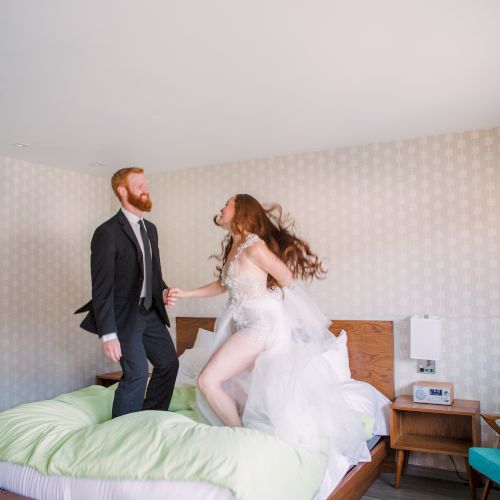 A couple, with the man in a suit and the woman in a white dress, are joyfully jumping on a bed in a light, modern bedroom.