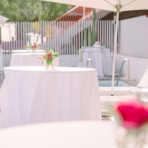 A poolside setup with white tablecloth-covered tables, each adorned with flower vases, alongside white chairs and umbrellas.