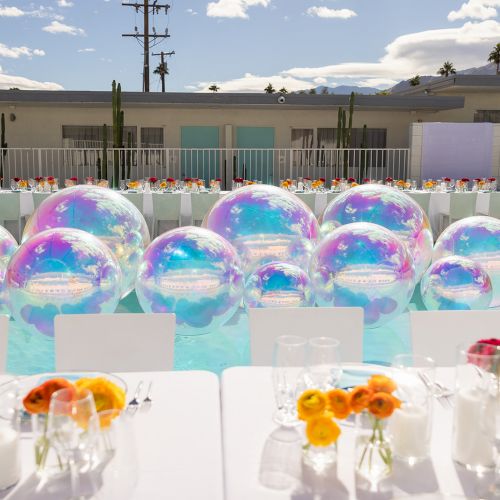 The image shows a poolside event setup with large iridescent beach balls in the water, surrounded by decorated tables and chairs.