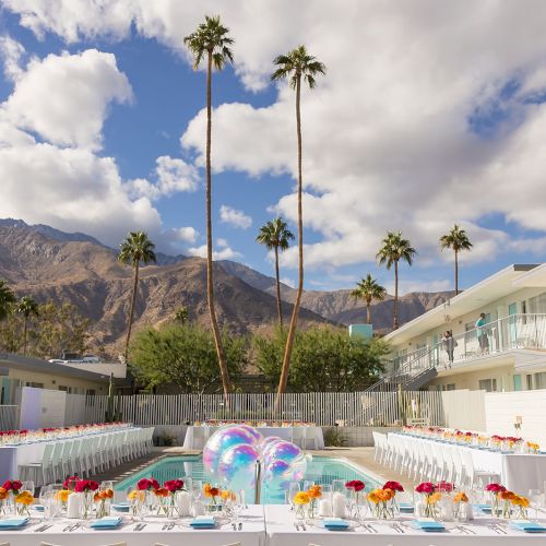 An outdoor event setup by a pool, with colorful table settings and mountains in the background, under a sunny sky with palm trees.