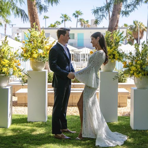 A couple stands facing each other, holding hands outdoors, surrounded by yellow flower arrangements, trees, and a building in the background.