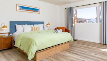 A neatly arranged bedroom features a bed with green bedding, a bedside table with lamps, an armchair, and a window with gray curtains.