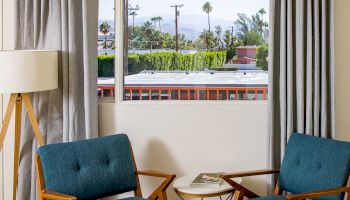 A cozy room with two blue chairs, a small white table, a floor lamp, and a large window with gray curtains, showcasing a view of palm trees outside.