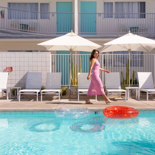 A woman in a pink dress walks by a pool with white lounge chairs and umbrellas, and colorful floaties are in the water.