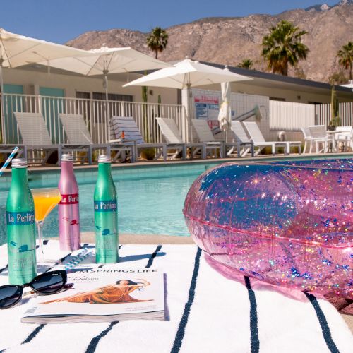 This image shows a poolside scene with beverages, a magazine, and a striped towel. A glittery inflatable is beside the towel.