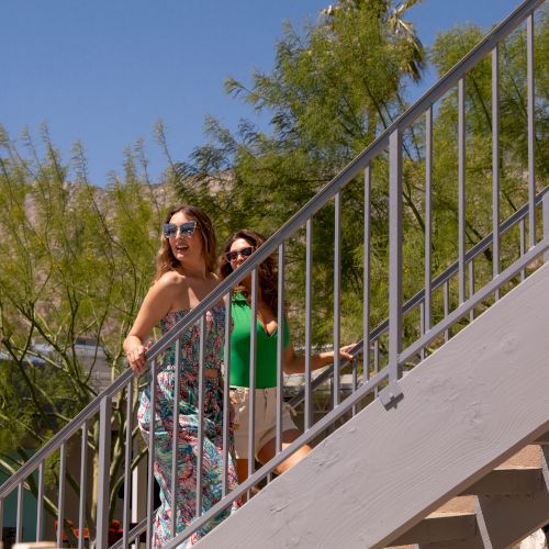 Two people are walking up an outdoor staircase on a sunny day, surrounded by green trees. They are dressed in summer attire and wearing sunglasses.