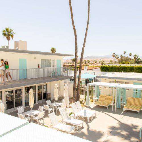 This image shows a bright, sunny scene at a small hotel or motel with a pool area, lounge chairs, cabanas, and a two-story building with guests.