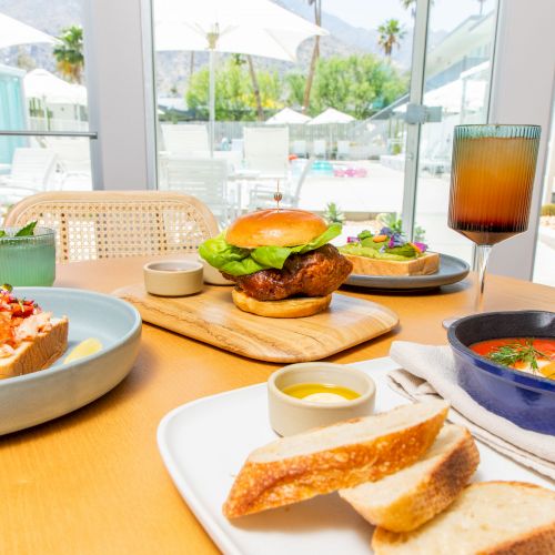 A table with a burger on a wooden board, a bowl of soup, toast, and a fruity toast dish, set in a bright, outdoor patio setting.