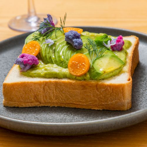 An open-faced sandwich topped with avocado slices, sliced cucumber, edible flowers, greens, and orange slices on a gray plate.