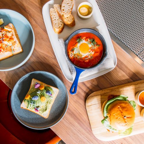 The image shows a variety of dishes on a table, including avocado toast, a burger with sauces, baked eggs in tomato sauce, and bread slices.