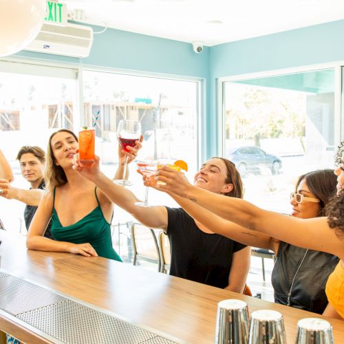 A group of people are gathered around a bar, raising their drinks in a toast, with smiles and laughter, enjoying a cheerful moment together.