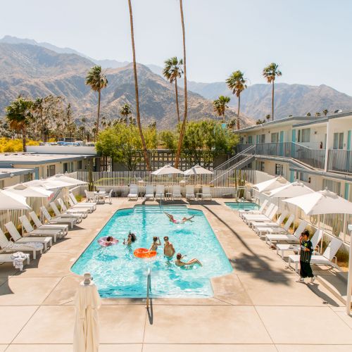 A pool surrounded by sun loungers and parasols at a motel, with people swimming and mountains in the background.