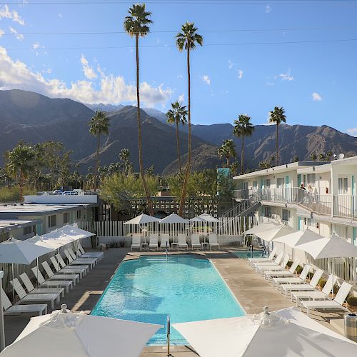 This image shows a hotel with a central outdoor swimming pool surrounded by lounge chairs and umbrellas, set against a backdrop of mountains and palm trees.