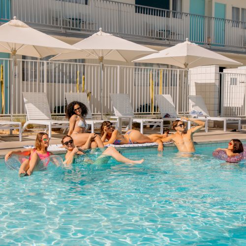 A group of people enjoying a sunny day by the pool, some in lounge chairs and others in the water with floaties, near umbrellas and a modern building.