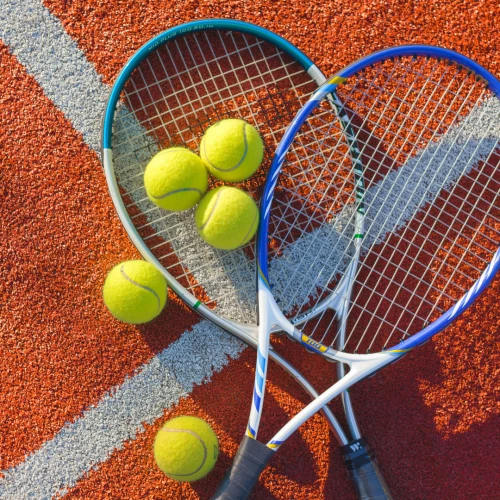 The image shows two tennis rackets and six yellow tennis balls on a red and white tennis court surface. The rackets are overlapping and crossed.