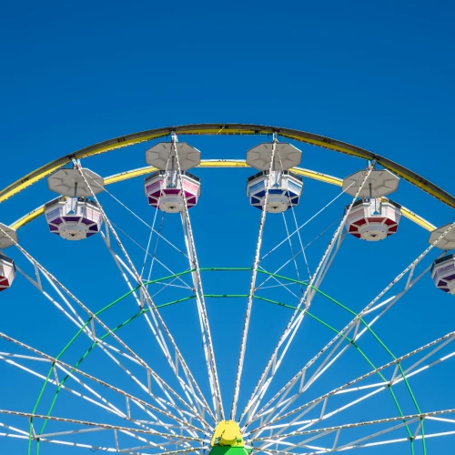 The image shows a Ferris wheel with colorful passenger cabins against a clear blue sky, capturing the top half of the attraction.