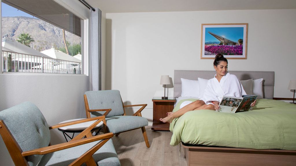 A person sits on a bed reading a magazine in a bright, modern room with two chairs near a window, and a framed picture on the wall.