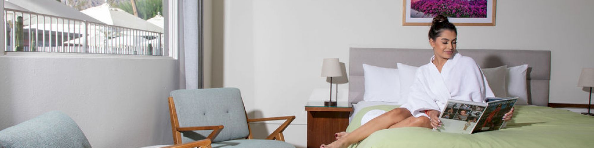 A person sits on a bed reading a magazine in a bright, modern room with two chairs near a window, and a framed picture on the wall.