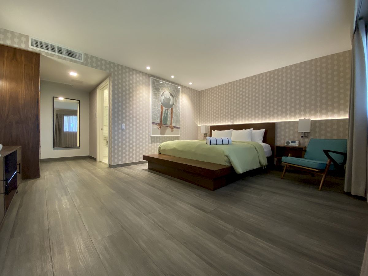 A modern hotel room with a large bed, stylish decor, and minimalistic furniture, featuring a wide view of the interior and ample hardwood floor space.