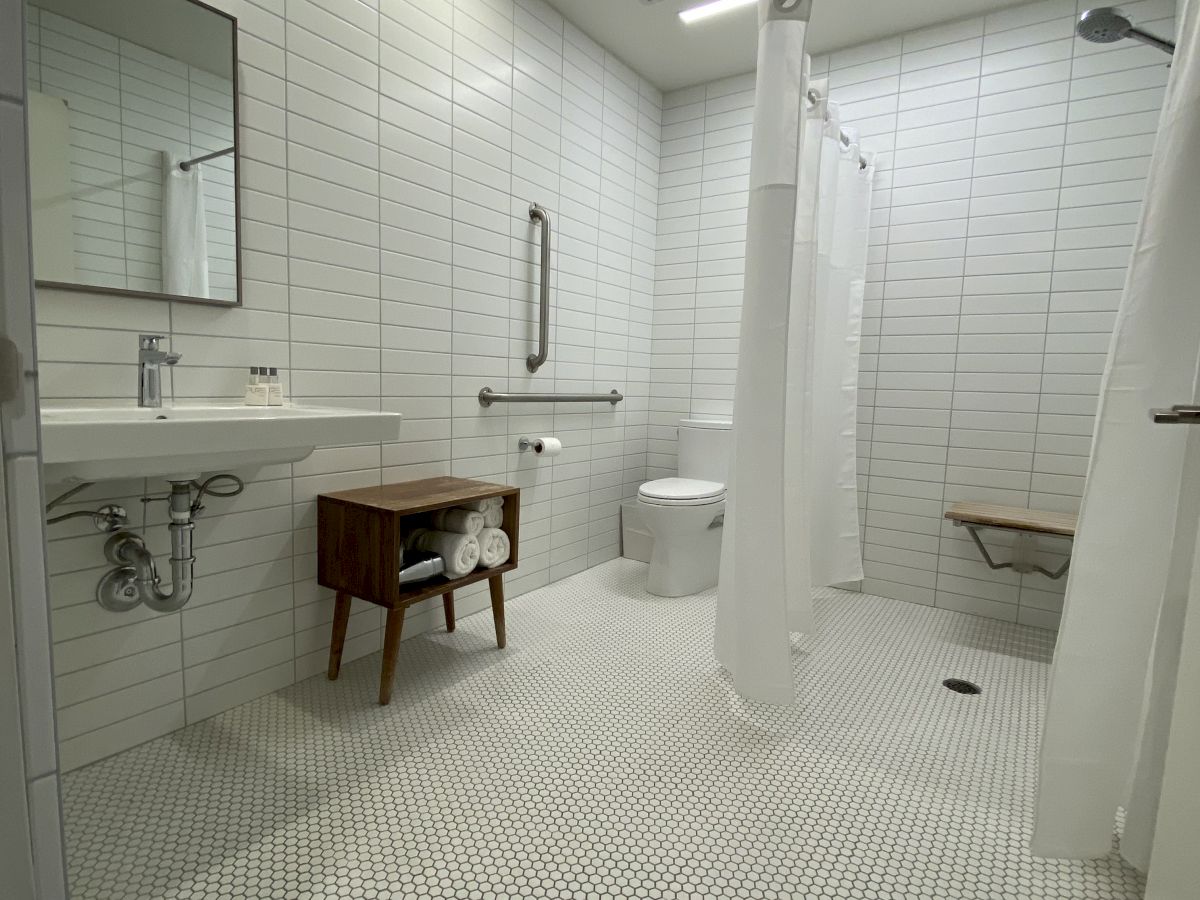 A modern bathroom with white tiles, a wall-mounted sink, a mirror, a small wooden cabinet, and an open shower area with a curtain and bench.