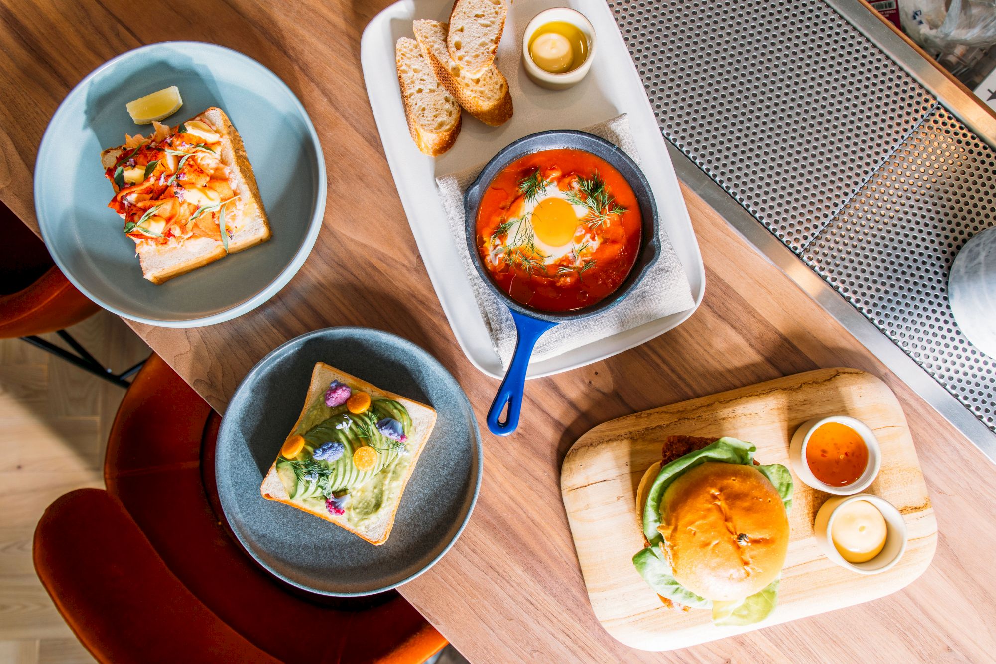 The image shows a table with various dishes, including avocado toast, a burger, crusty bread with a small dish of soup, and a skillet meal.