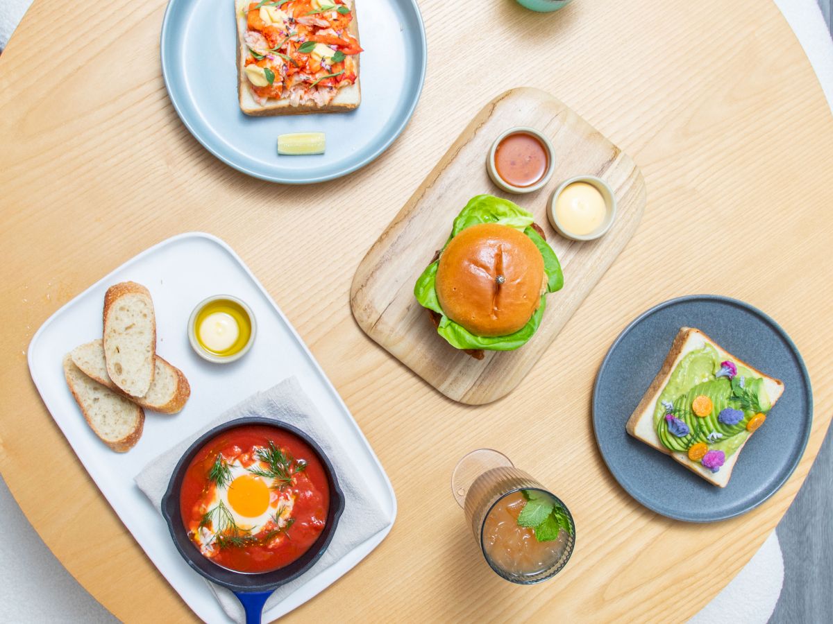 The image shows a table with a variety of dishes, including a burger, toast with toppings, a skillet dish with an egg, and two drinks.