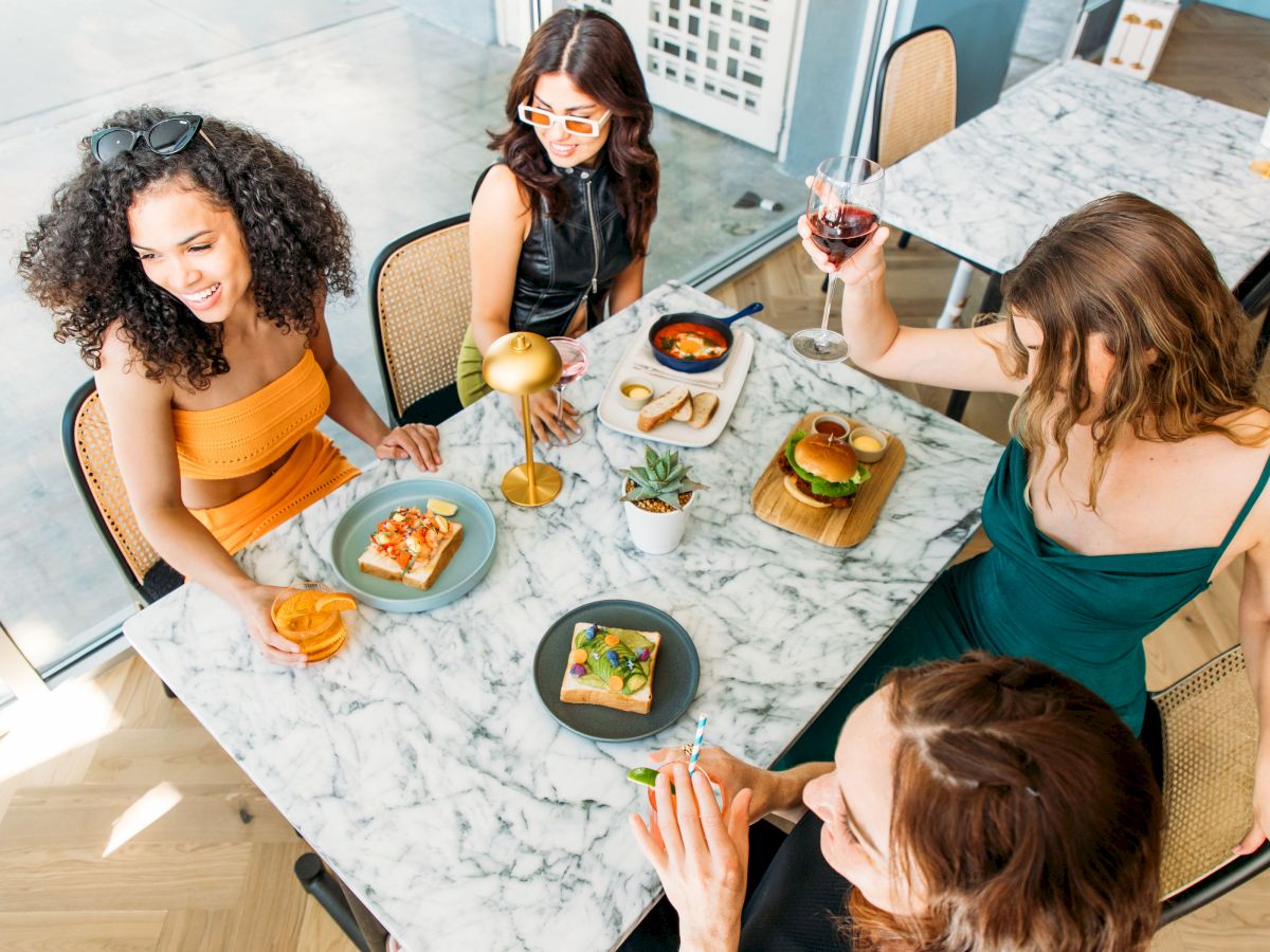 Four women are seated around a table with various dishes and drinks, enjoying a meal together in a brightly lit restaurant.