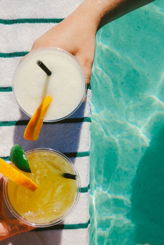 Two hands holding drinks by a pool, with a striped towel underneath. The sky is reflected in the water, giving a relaxing summer vibe.