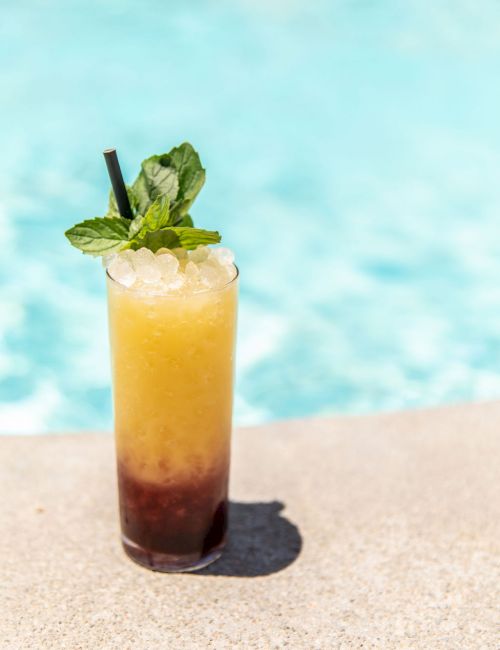 A refreshing cocktail garnished with mint leaves and a straw, placed beside a swimming pool in bright sunlight.