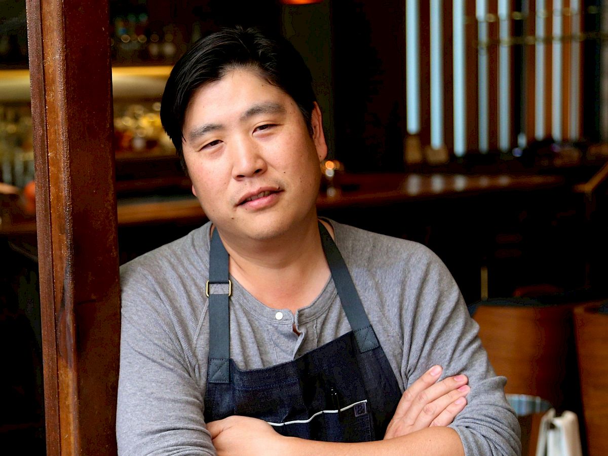 A person in a gray shirt and black apron leans against a wooden beam in a dimly lit setting, arms crossed, with a restaurant background, ending the sentence.